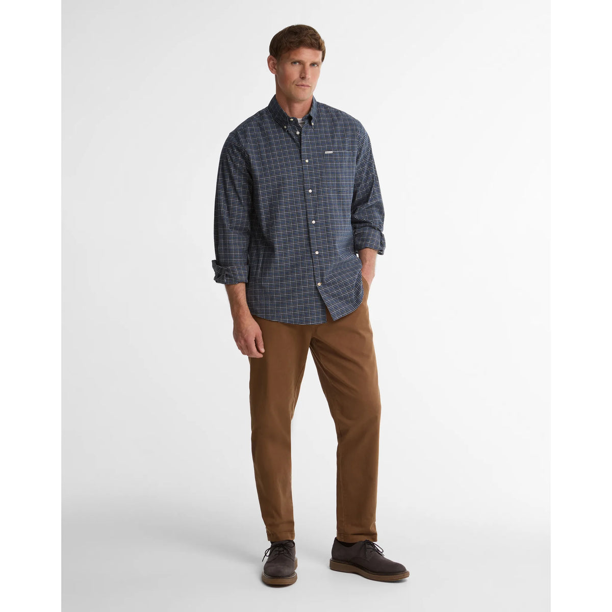 A man modeling a long-sleeve button-down shirt with a dark blue and gray micro-check pattern, paired with brown trousers and dark brown shoes, against a light gray studio background.
