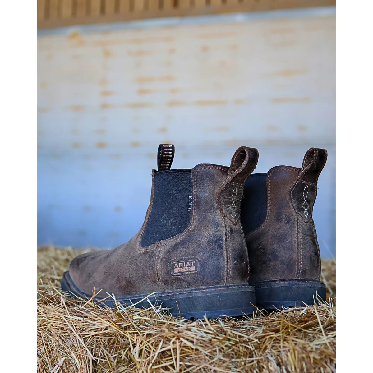 A pair of Ariat dark brown leather Chelsea boots resting on a bed of straw hay with a rustic wooden wall in the background.
